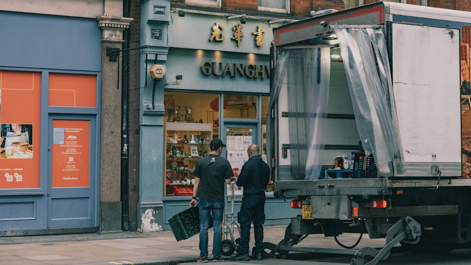 Two men are standing on a sidewalk in front of a storefront with Asian signage and the word 'GUANGH' visible above the entrance. One man is holding a black plastic crate, while the other appears to be pointing or giving instructions. A moving truck with an open rear door and a plastic curtain is parked adjacent to the storefront, prepared for furniture and box loading as part of a house removal or relocation process. The moving vehicle is positioned partially on the pavement, with visible equipment such as a lifting frame or trolley nearby. The setting suggests the loading or unloading phase of packing and moving services, with the transfer of packed boxes, furniture, or appliances between the truck and the property. The scene is outdoors, well-lit, with the building’s detailed features, including signage and window displays, clearly visible, reflecting professional movers engaged in a home relocation in an urban environment.