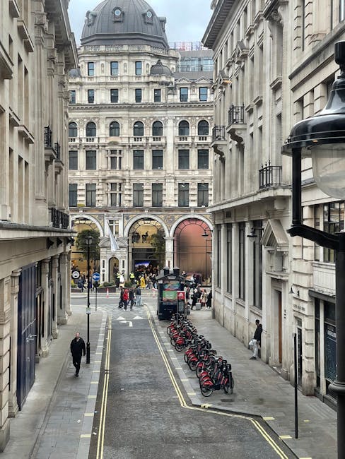 A historic stone building with ornate architectural details, including sculpted figures and decorative columns, is situated on a city street in London near Aldwych. The building has large arched windows with dark frames, some of which are partially obscured by parked vehicles. In the foreground, a pavement with light-colored stone slabs and tactile paving for accessibility is visible. The street features traffic lights and bollards, and a small portion of a moving vehicle can be seen on the left. The environment is urban, with modern high-rise buildings visible behind the historic structure. This scene relates to house removals and moving services provided by Man with Van Aldwych, highlighting the logistical challenges of furniture transport and home relocation in central London.