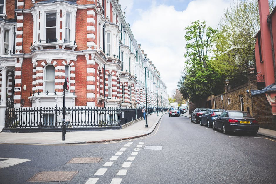 A residential street scene taken during daylight hours, showing a row of white, multi-story Victorian-style terraced houses with decorative window frames, balconies, and some small front gardens enclo