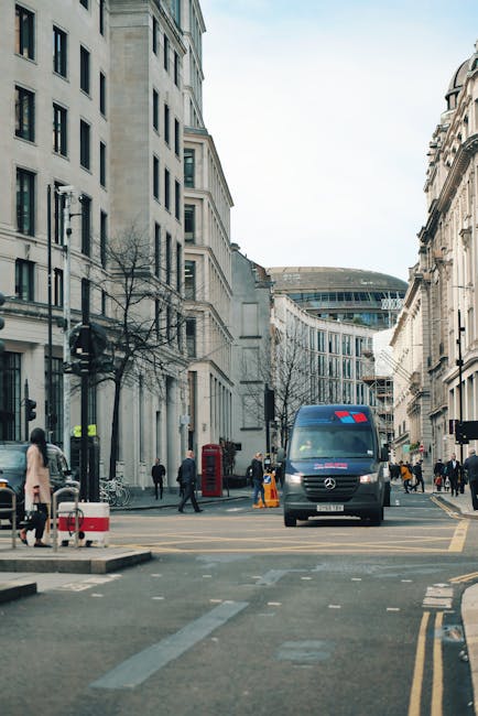 A delivery van parked on a busy city street in Aldwych, with the rear doors open for a home relocation. Inside the van, visible packing materials such as cardboard boxes, bubble wrap, and protective blankets are prepared for furniture transport. Several people are walking nearby, some approaching the van, which is positioned close to the pavement to facilitate loading. The surrounding environment includes classic London-style buildings with multiple windows and a mixture of modern and traditional architecture. Street furniture, including a red telephone box, is present on the sidewalk, while the road markings indicate a controlled loading zone. The presence of a trolley or straps is not visible, but the scene suggests an organized moving process with crew members from Man with Van Aldwych ready to assist in a house removals or furniture transport operation, ensuring efficient packing and loading for a house move in the Aldwych area.