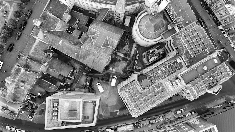 An aerial black-and-white photograph depicts a narrow urban alleyway surrounded by several multi-story buildings, some with flat roofs and others with pitched roofs covered in roofing materials. The alley contains a few parked cars, mostly small vans and sedans, positioned close to the buildings' entrances. There are visible footprints and minor debris scattered across the ground, indicating activity. Partially inside the alley, a Man with Van Aldwych crew is in the process of loading or unloading furniture and boxes, which are wrapped in plastic and placed on dollies or carts. A small stack of stacked cardboard boxes and padded blankets lie nearby, ready for transport. The scene captures the busy loading process typical of home relocation or furniture transport services, with the crew actively managing the belongings for a prompt move through the limited access area. Bright lighting highlights the details of the objects and the environment, emphasizing the logistical effort involved in house removals in a dense city setting.