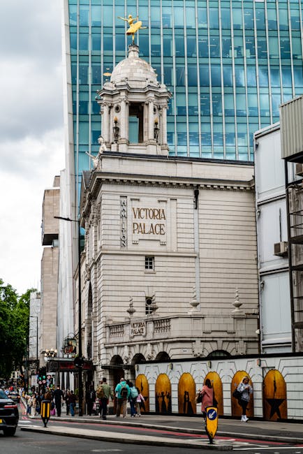 The image shows the Victoria Palace Theatre in Aldwych, London, featuring a historical facade with ornate architectural details including classical columns, decorative sculptures, and a domed tower to