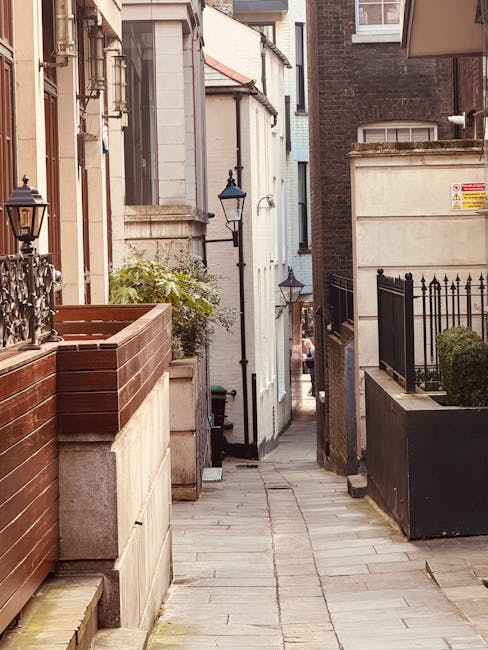 A narrow urban alleyway with a paved stone pathway flanked by residential buildings on both sides. On the left, there is a wooden planter box atop a concrete wall, holding some greenery, and a decorative black lantern mounted on the wall. The building facades vary from light-colored brick to darker brickwork; some windows and doors are visible, along with external fixtures like a street lamp attached to one building. On the right, a black metal fence borders a small garden space with bushes, and a large black container or planter is positioned near the entrance. At the far end of the alley, a person is partially visible, carrying or moving items, consistent with a home relocation or furniture transport process managed by Man with Van Aldwych. The overall scene reflects a typical city setting with limited access pathways, emphasizing the logistical challenges of staircase, narrow mews, and lift access during removals or moving services.