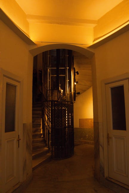 Indoor view of a narrow staircase and mews access area inside a building, with a cylindrical lift shaft visible at the top of the stairs. The space features a curved ceiling and walls painted in light cream tones, with two white doors on either side of the entrance. The staircase is made of wood with a dark metal railing, positioned next to the lift shaft, which has a protective metal mesh surrounding it. The area is illuminated with warm lighting, highlighting the confined space typical of a city mews residence. This image illustrates the challenges of home relocation in tight spaces, where careful furniture transport and packing are necessary, as seen in the context of a house removals service like Man with Van Aldwych.