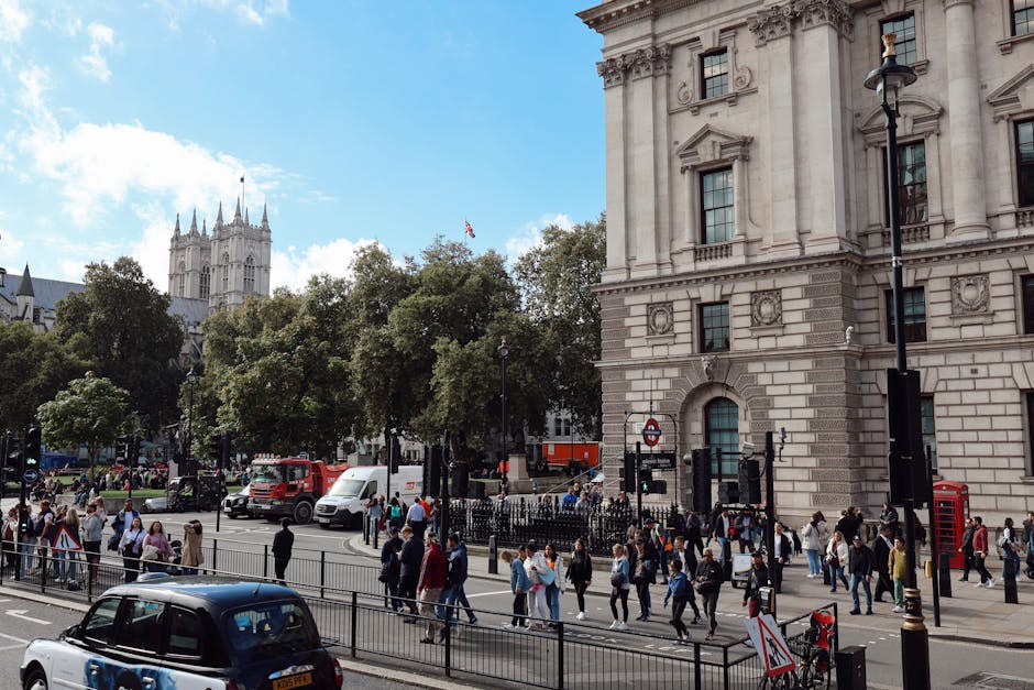 A busy street scene in Aldwych, London, with pedestrians walking on the sidewalk and crossing at a designated crossing, in front of a large historic stone building featuring detailed architectural ele